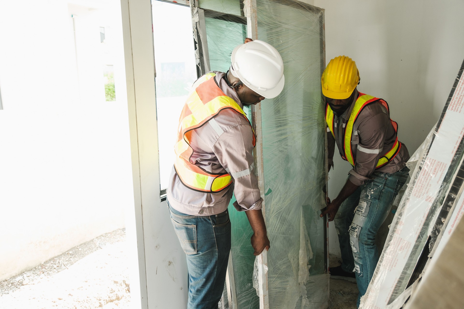 Two construction workers carefully install a large glass panel in an indoor site. Wearing safety helmets and vests, they collaborate on precise handling, showcasing modern building teamwork.